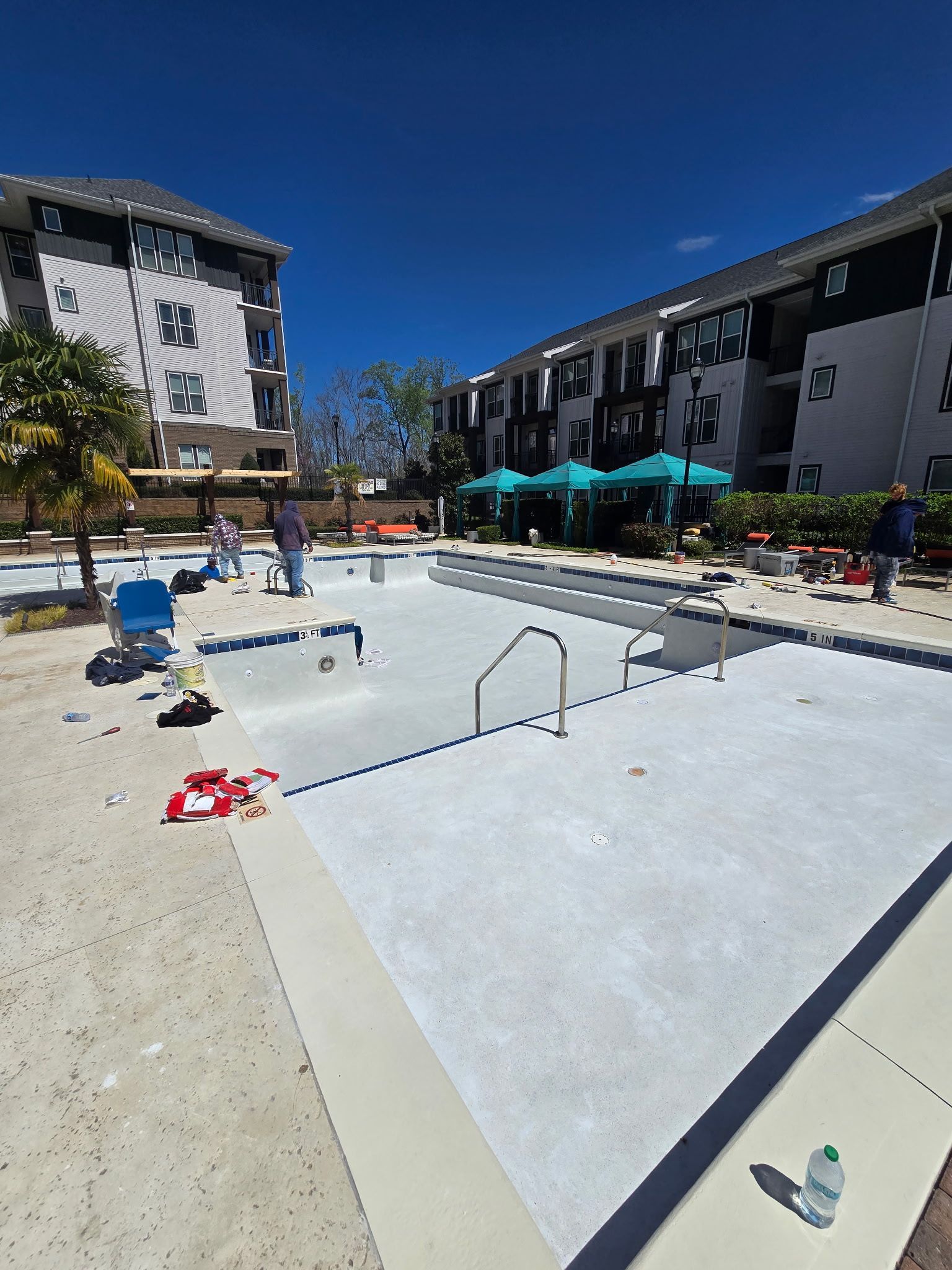 Outdoor apartment pool under construction, with white plaster, ladders, and lounge chairs under a bright blue sky
