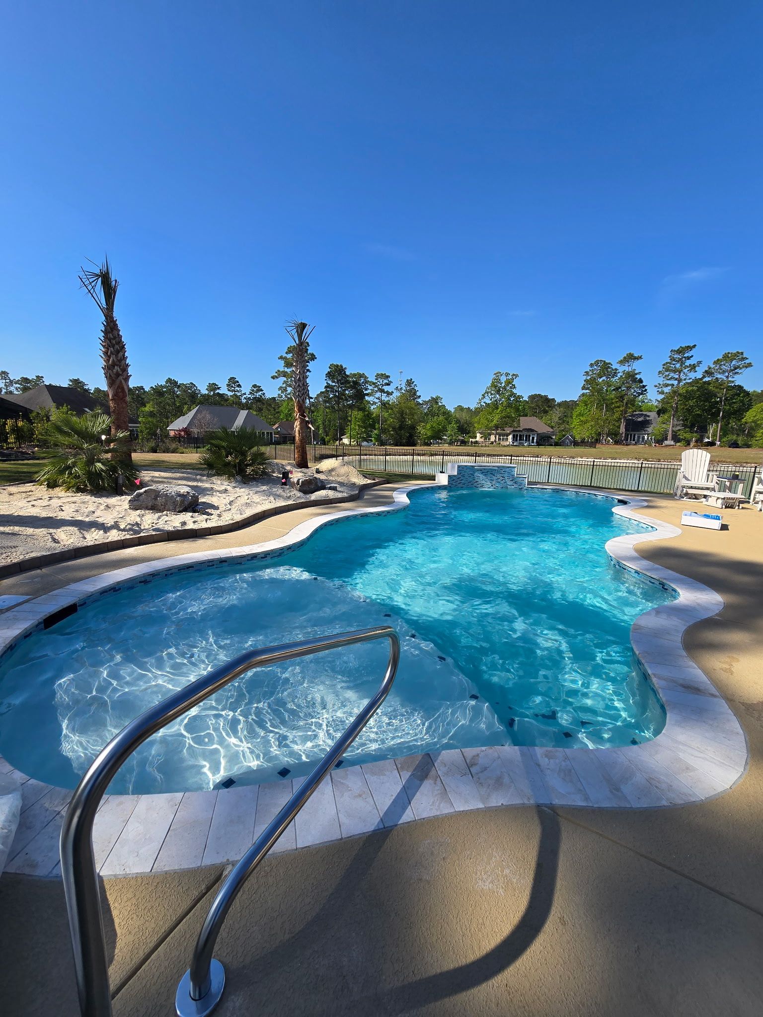 Curved resort pool with blue water, stainless handrail, lounge chairs, and palm trees under a clear sky