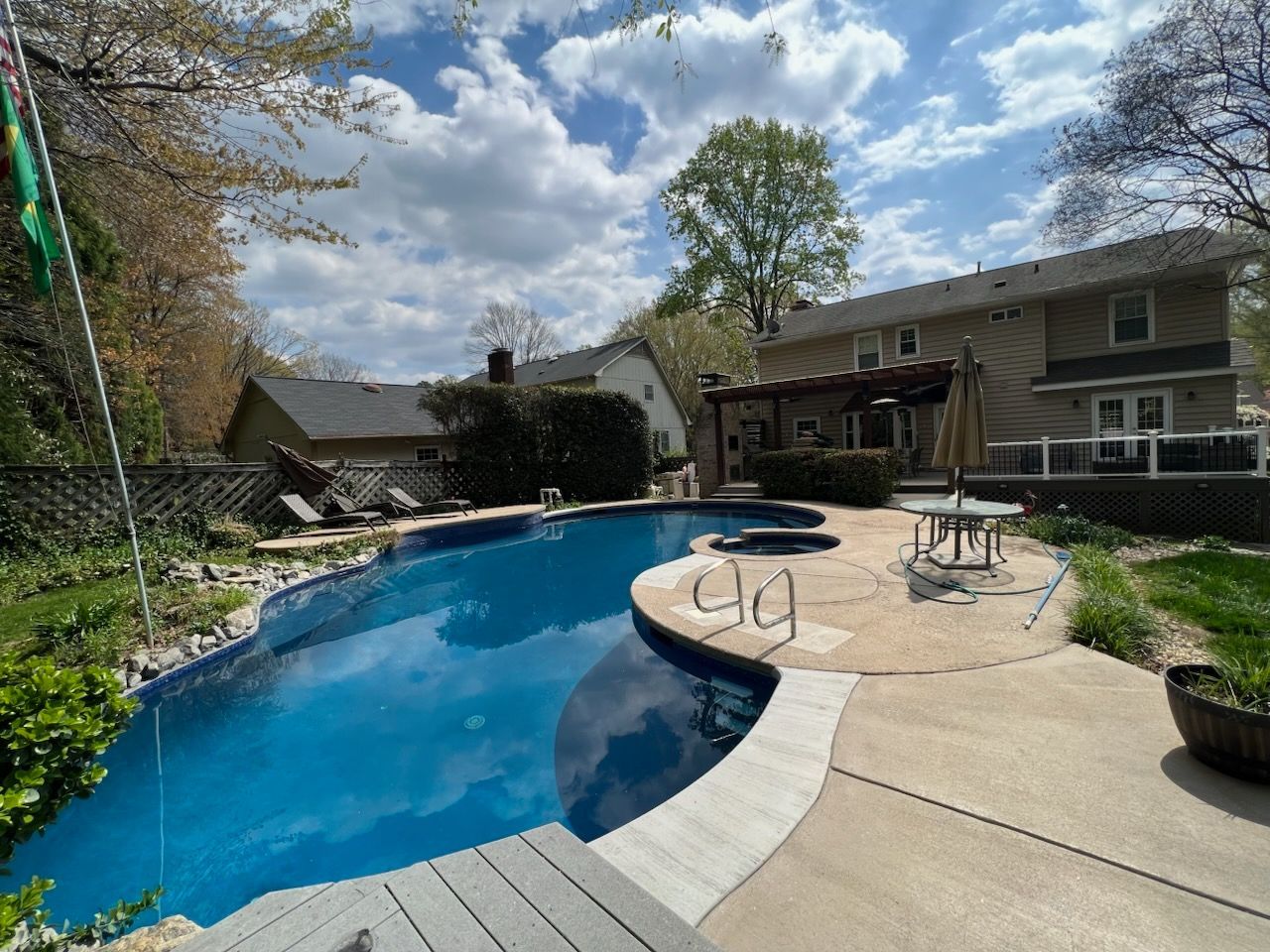 Backyard pool with curved blue water, patio seating, and a house in the background under a cloudy sky