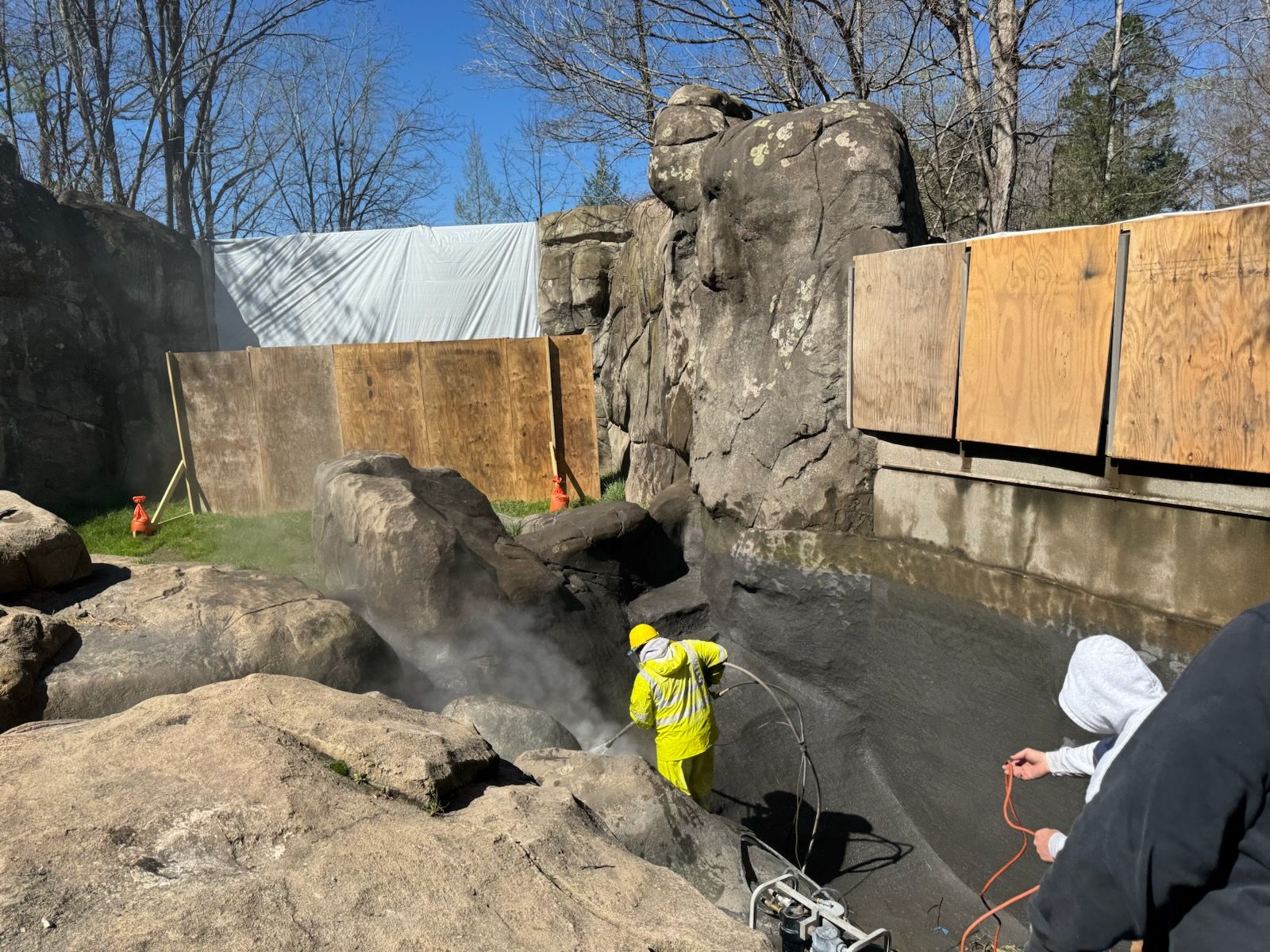 Workers in neon safety gear spray water near large rocks at a zoo enclosure under construction.