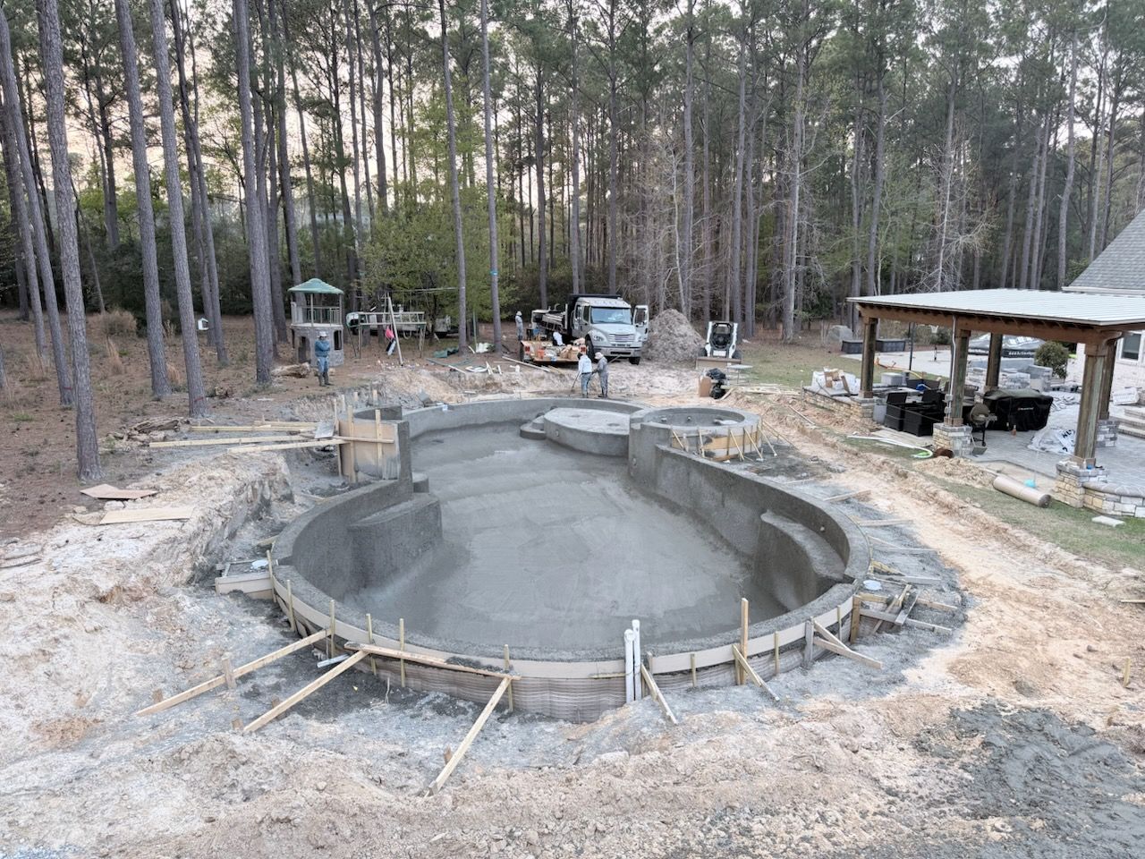 Large unfinished concrete pool or pond in a wooded yard, with construction materials and a small shelter nearby