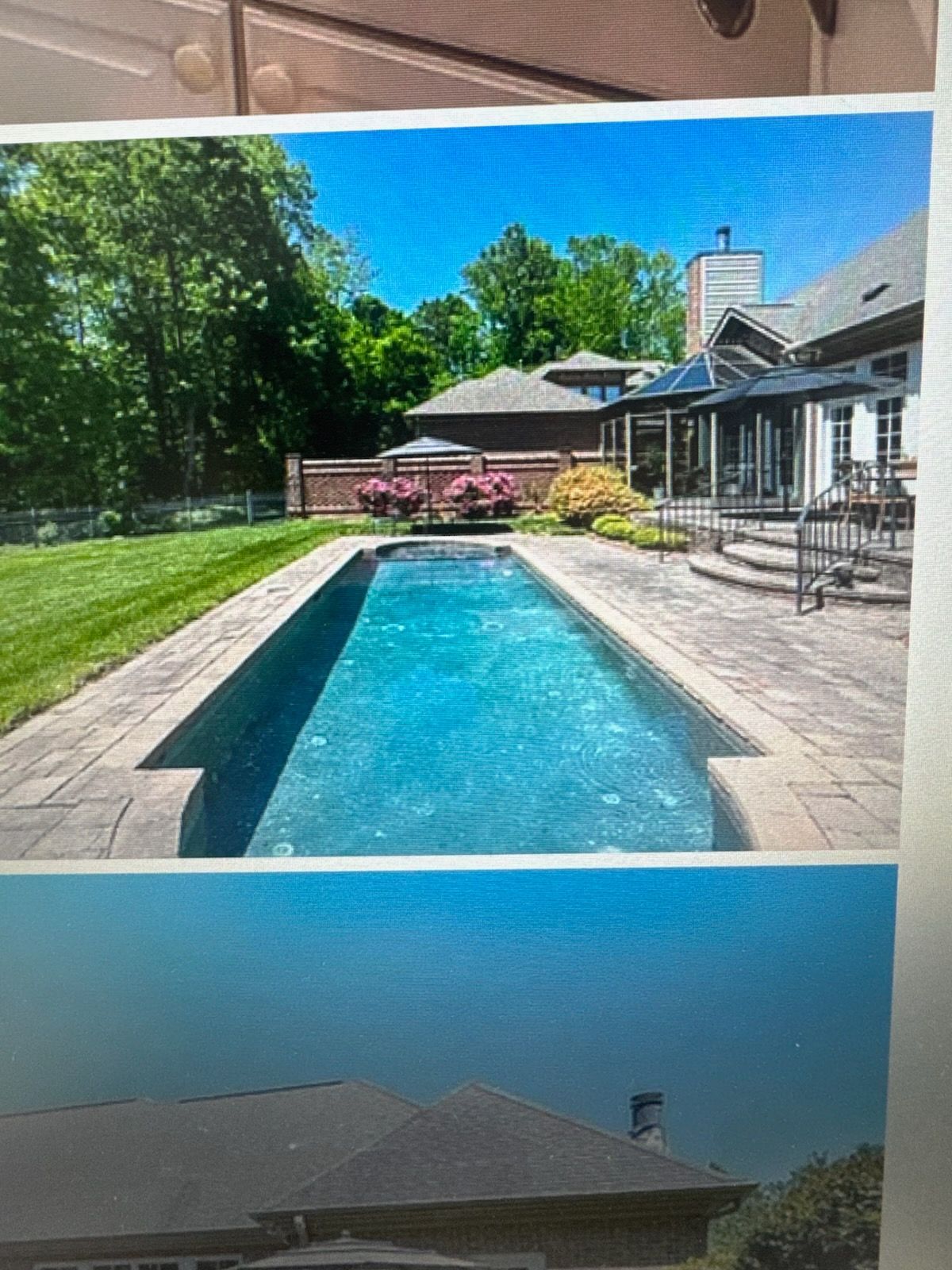 Backyard with narrow blue pool, green lawn, patio chairs, and a house under a clear sky.