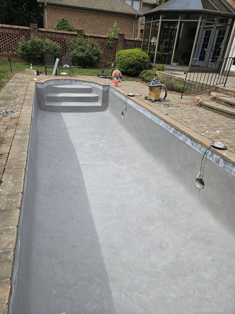 Empty concrete backyard pool under construction beside a brick house and patio.