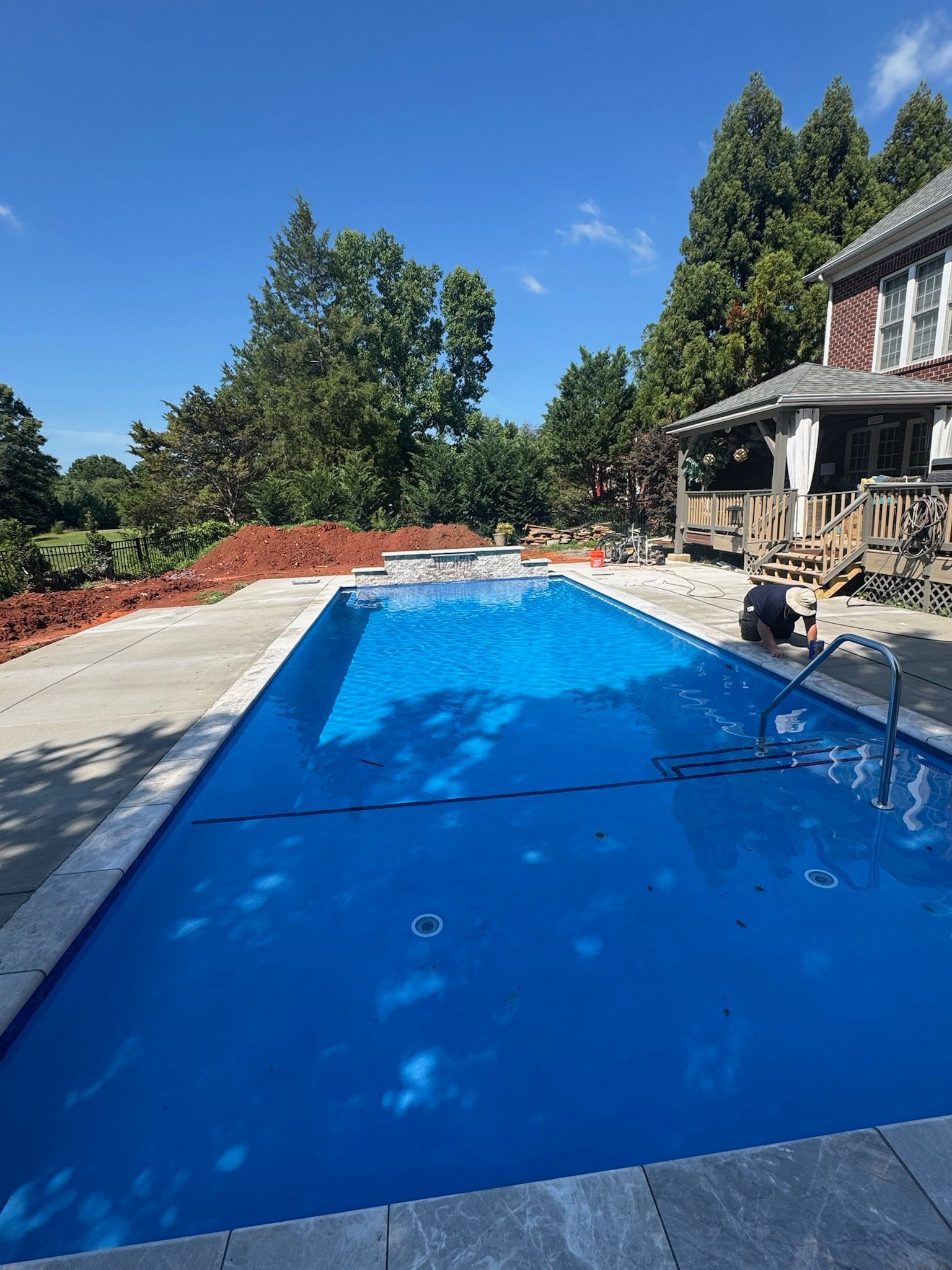 Blue swimming pool beside a stone patio and house, with trees and a clear sunny sky