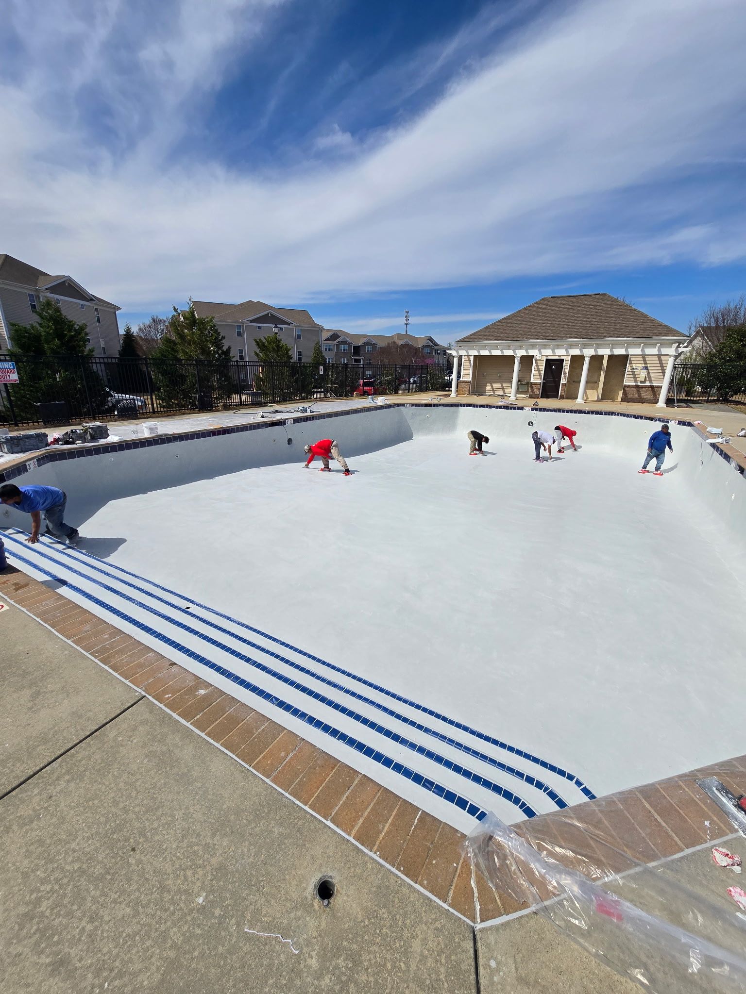Outdoor empty swimming pool with people playing in a white bowl, under a blue sky and nearby houses.
