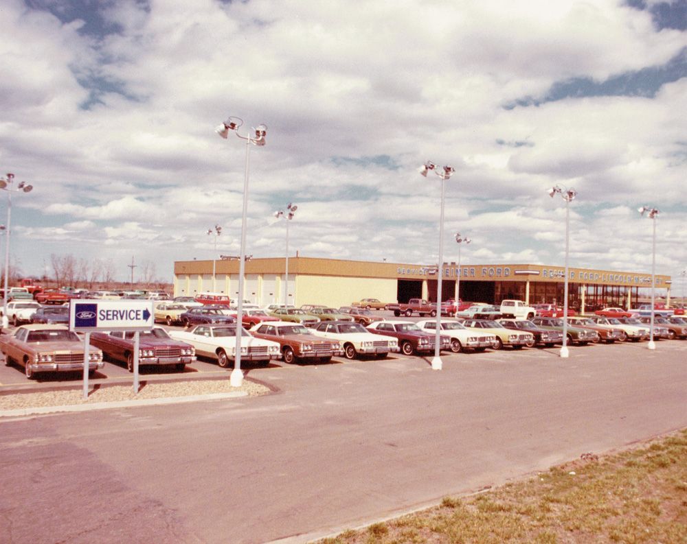 Car dealership with rows of cars, service sign, and building under cloudy sky.