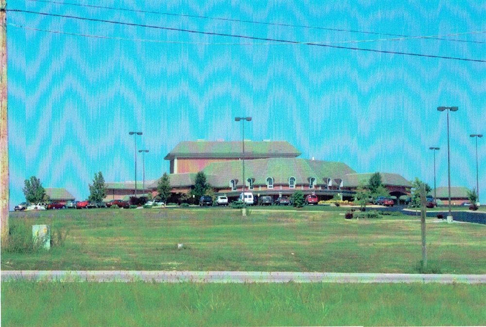 Building with red brick and green roof on a grassy hill, under a wavy blue sky.