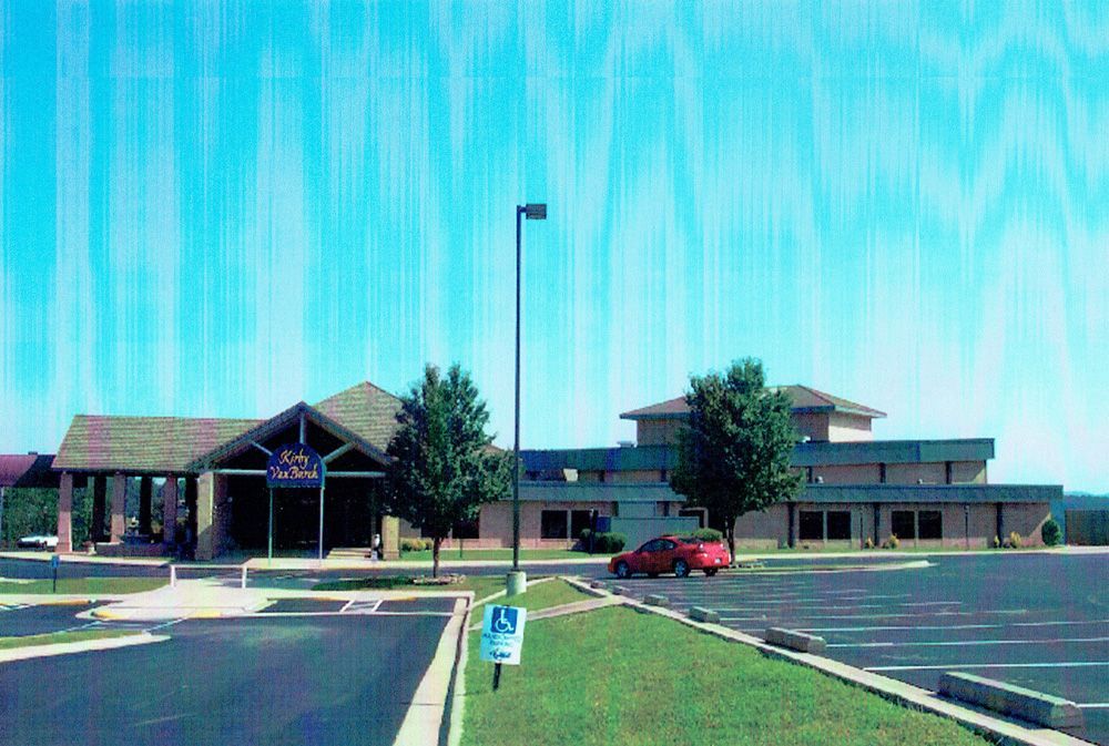 Building with a covered entrance, parking lot, red car, and blue sky.