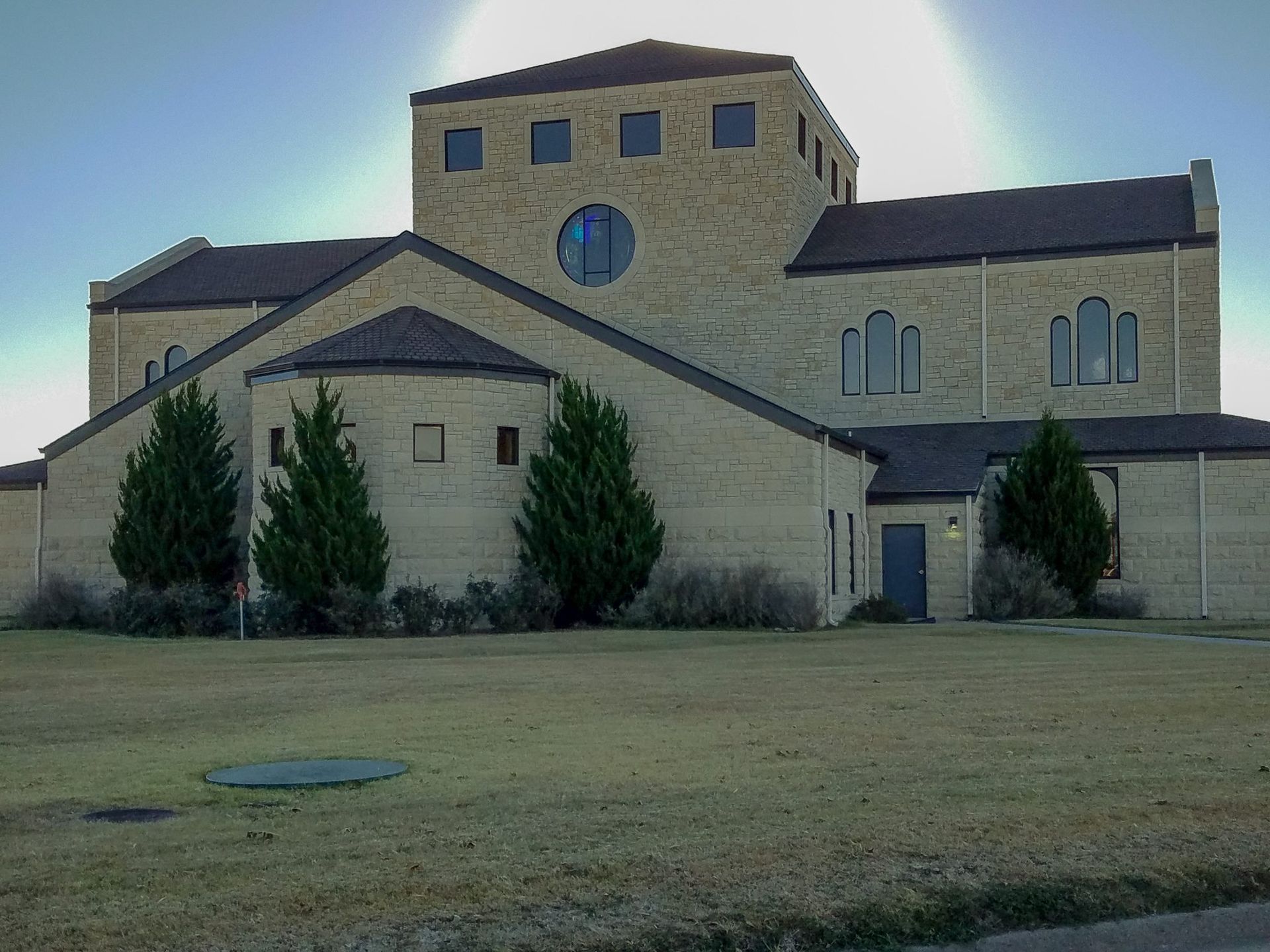 Tan brick church with multiple gabled roofs, a round window, and evergreen trees on a grassy lawn.