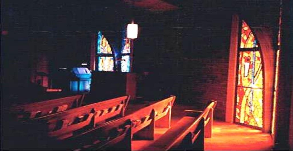 Dark church interior with sunlight streaming through stained glass windows onto wooden pews.