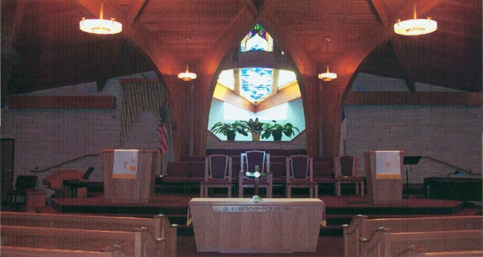 Interior view of a church sanctuary with wooden arches, altar, stained glass window, and rows of pews.
