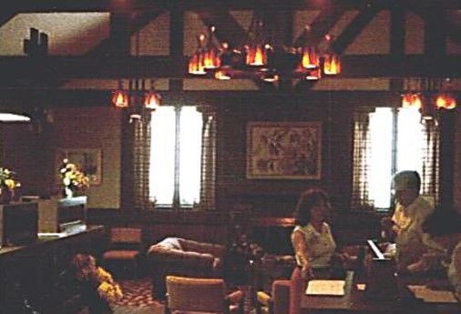 Interior of a dimly lit room with people. Dark wood paneling, windows with blinds, and a chandelier are visible.
