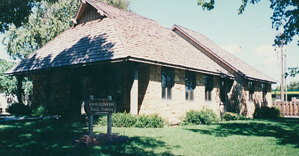Stone building with a shingled roof and small windows, set on a green lawn with a sign.