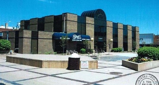 Brick building with blue awning and glass windows on a paved plaza under a clear sky.