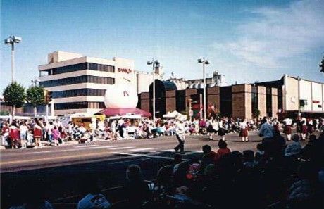 Parade on a city street. Spectators watch as performers march past buildings, including a multi-story office building.