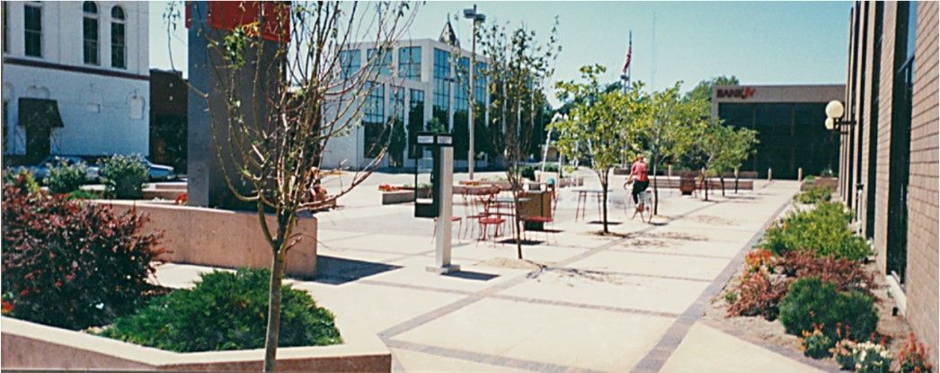 Outdoor plaza with trees, plants, and seating areas. A person walks by trees in the distance.