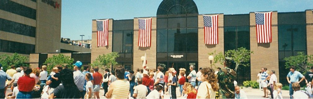 Large crowd in front of a building decorated with American flags. A tall building is to the left.