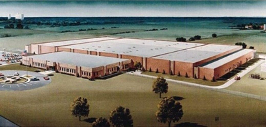 An industrial complex with multiple buildings, set in a green field with trees, and a blue sky in the background.