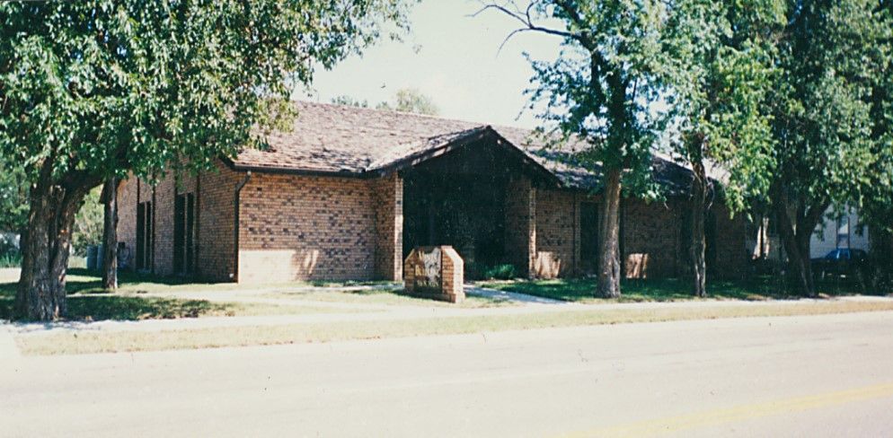 A brick building with a brown roof, flanked by trees, sits behind a street.