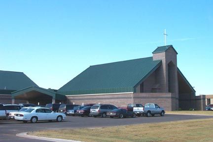 Church building with green metal roof, cross on top, and cars parked in front.