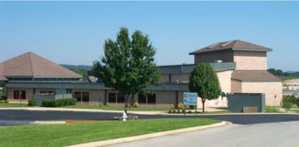 A light brick building with a brown roof on a sunny day. A tree and grass are in the foreground.