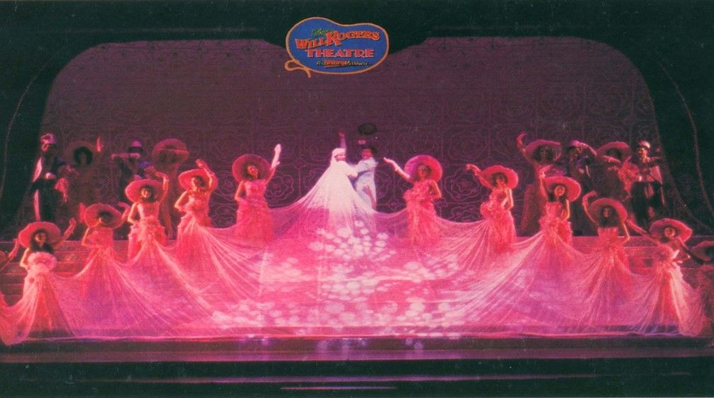 Stage production with dancers in pink dresses and hats, a couple in white, under a blue sign.