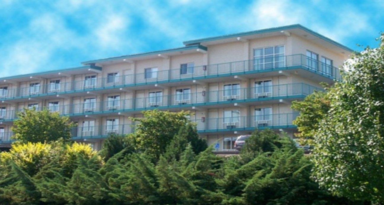 Multi-story building with balconies, set among trees against a blue sky.