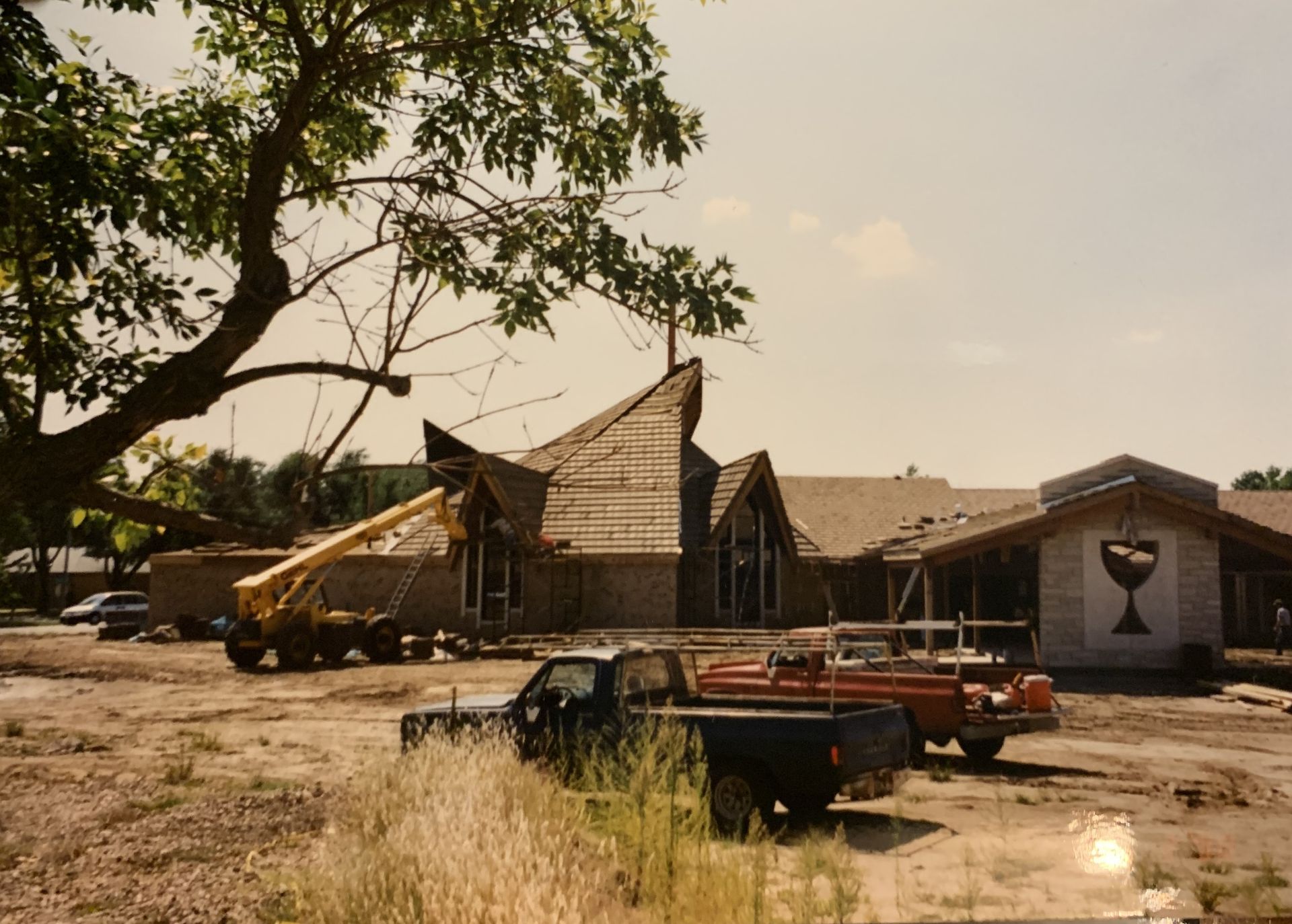 Church construction site; wooden framework, crane, trucks, brown dirt ground, sunny day.