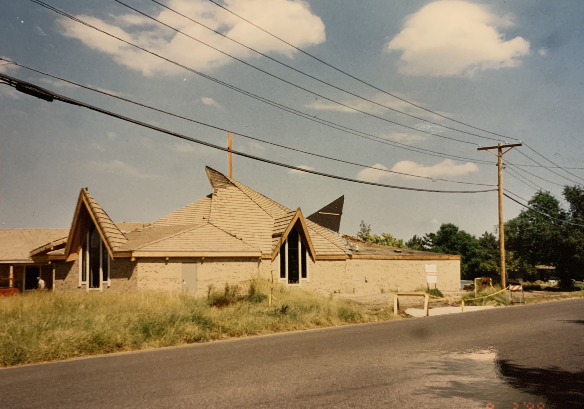 A building with unique rooflines, overgrown grass, power lines overhead, and a road.