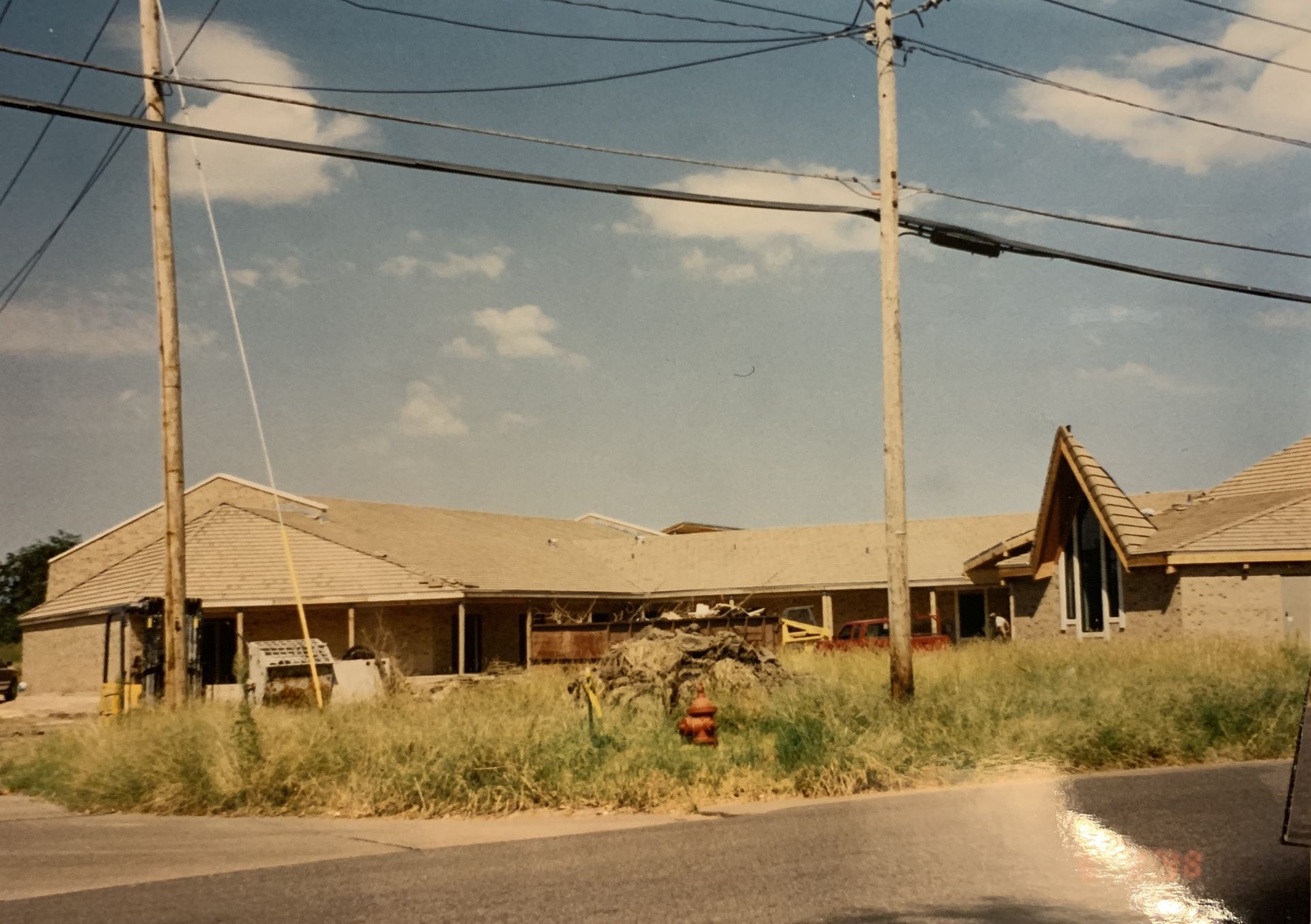 Building under construction with overgrown grass, telephone poles, and power lines against a cloudy sky.