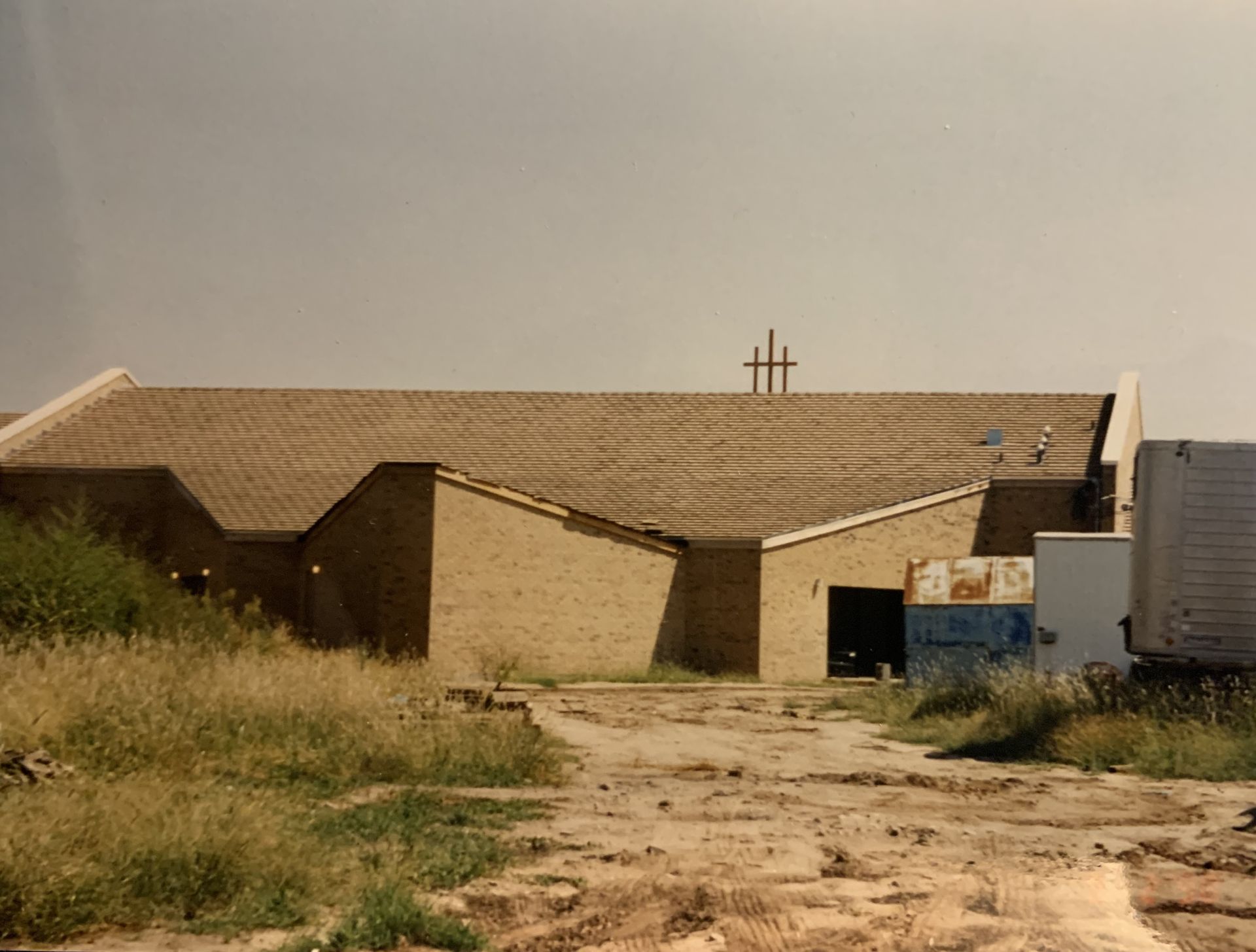 Brick building with a sloped roof and a cross on top, dirt path in the foreground, trailer on the right, cloudy sky.