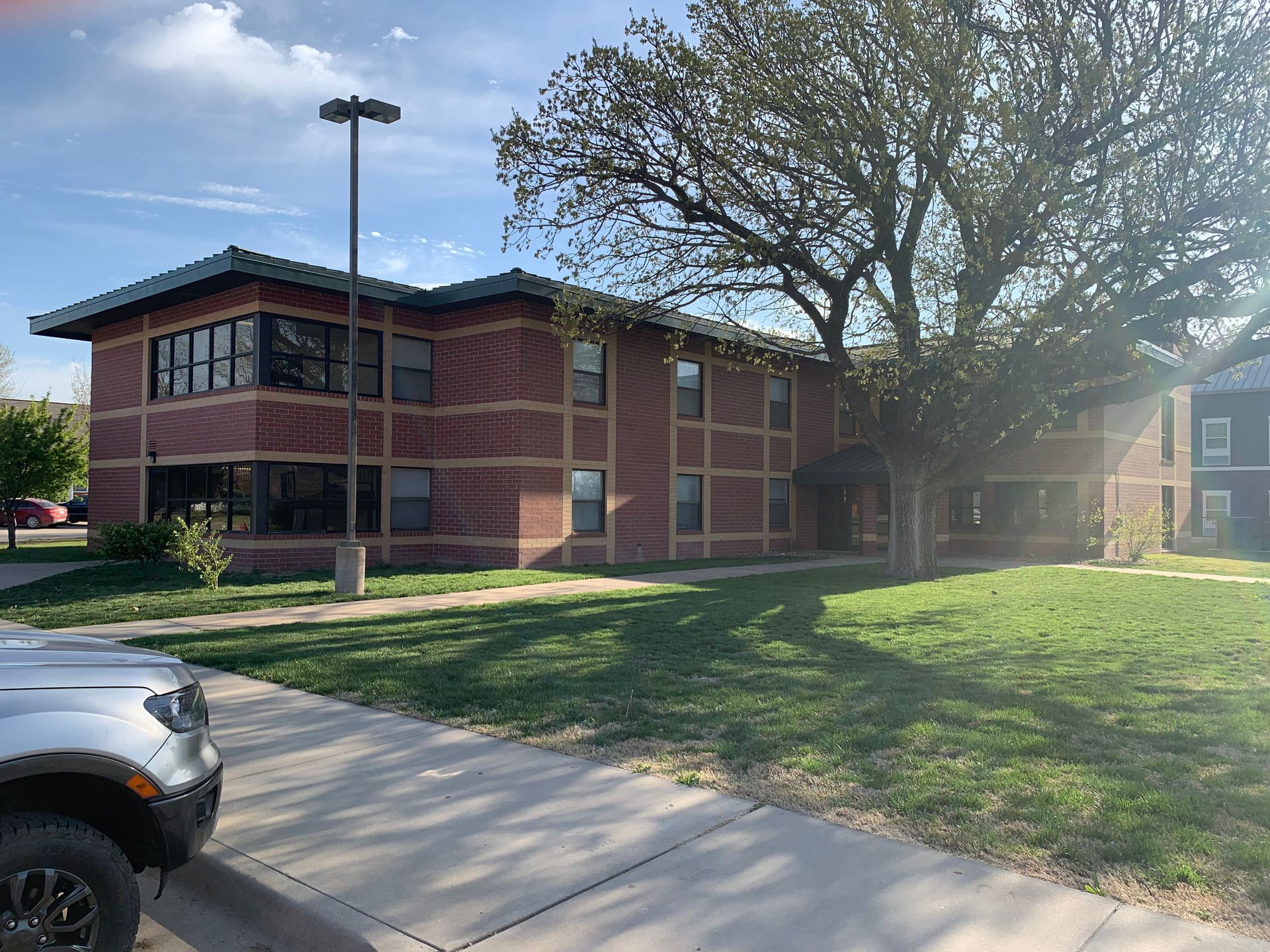 Two-story brick building with large windows and a green lawn. A tree provides shade. A silver vehicle is parked in front.
