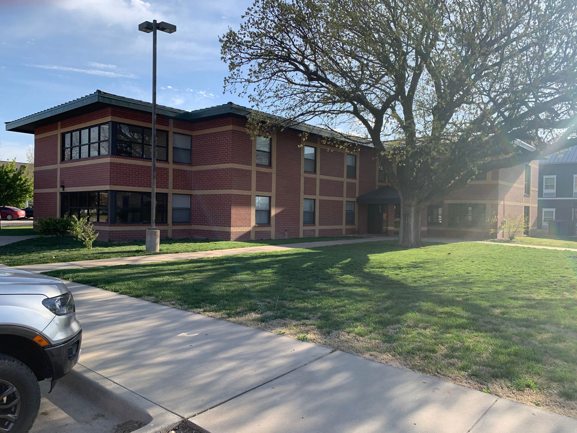 Brick two-story building with many windows, a grassy lawn, and a large tree; a gray vehicle is parked in the foreground.
