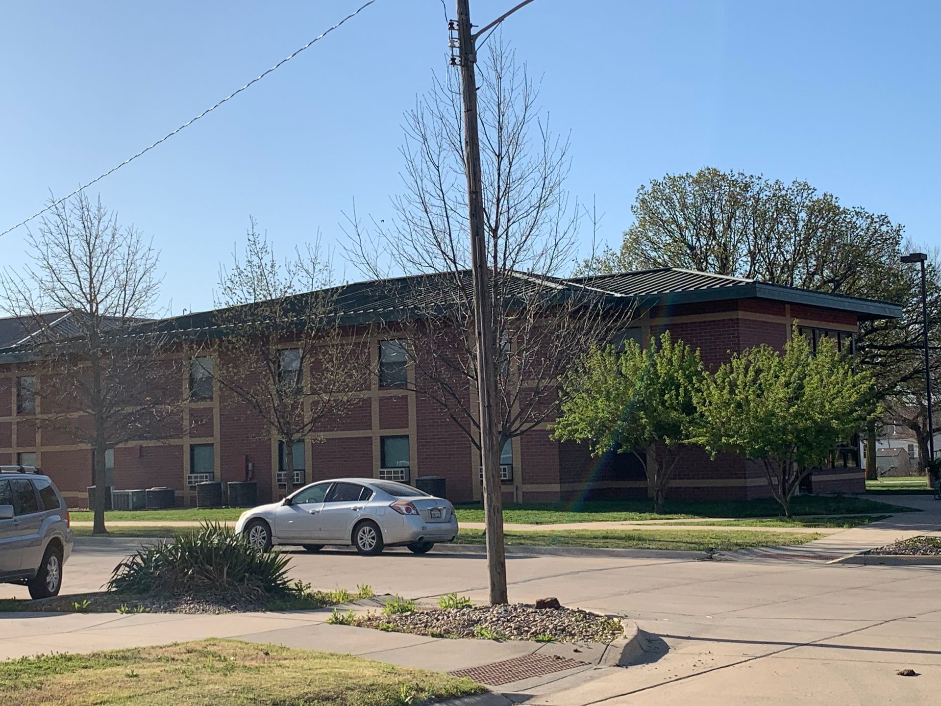 Brick building with windows, a silver car parked on street, trees, and a clear sky.