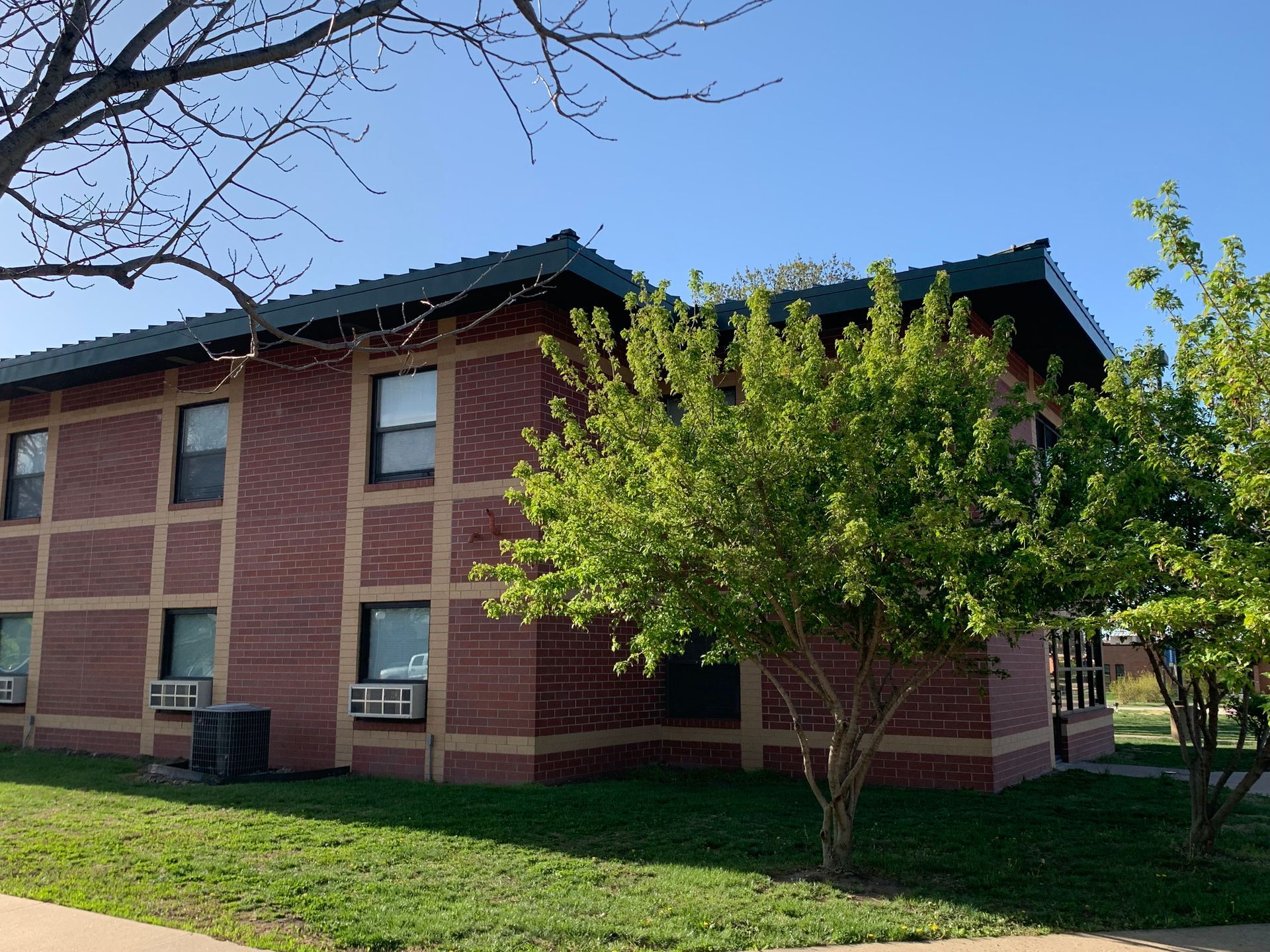 Two-story brick building with green roof, windows, and trees on a sunny day.