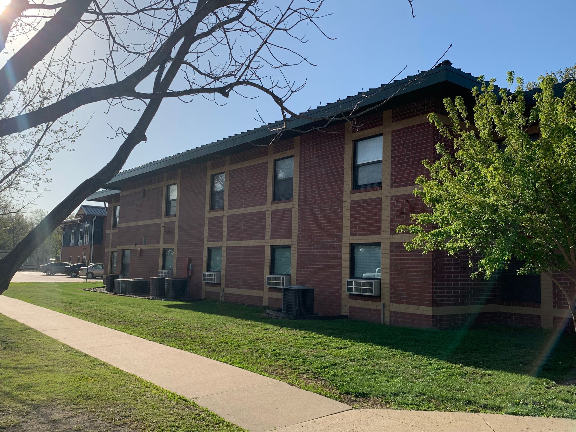 Two-story brick apartment building with green trim and air conditioning units, sidewalk, and trees.