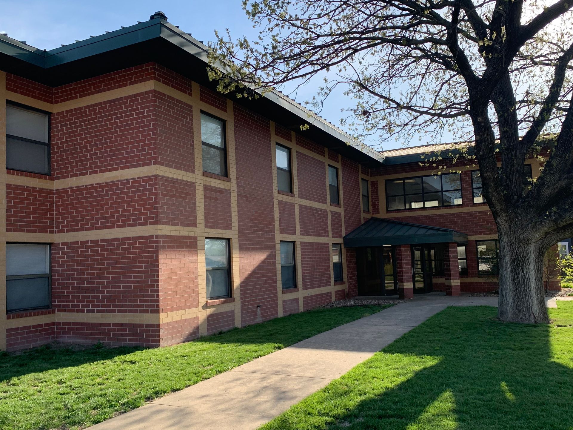 Two-story brick building with green roof trim and a walkway leading to a glass door under a small canopy, tree on the right.