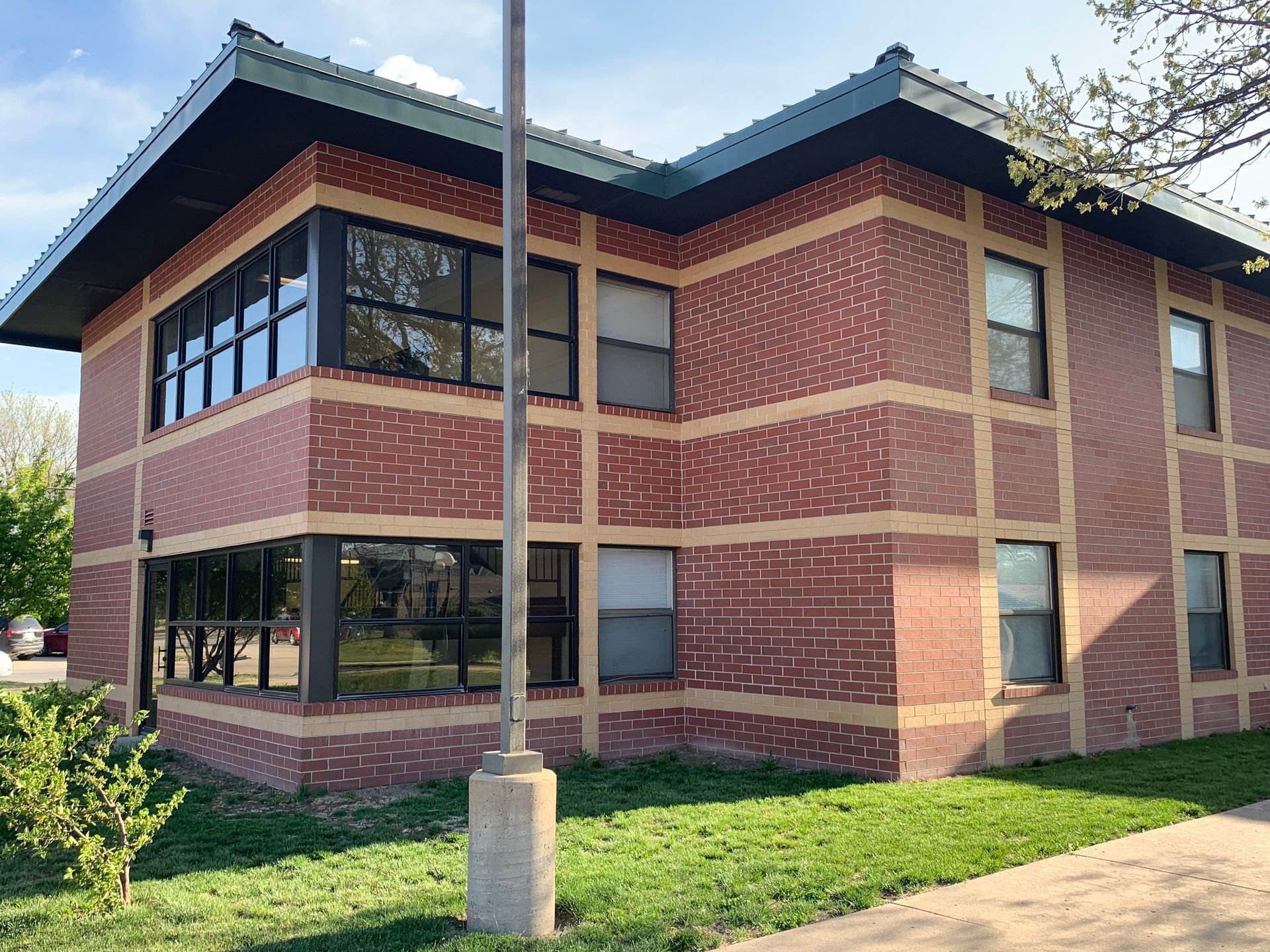 Two-story brick building with large windows and a green roof, on a grassy lawn with a flagpole.