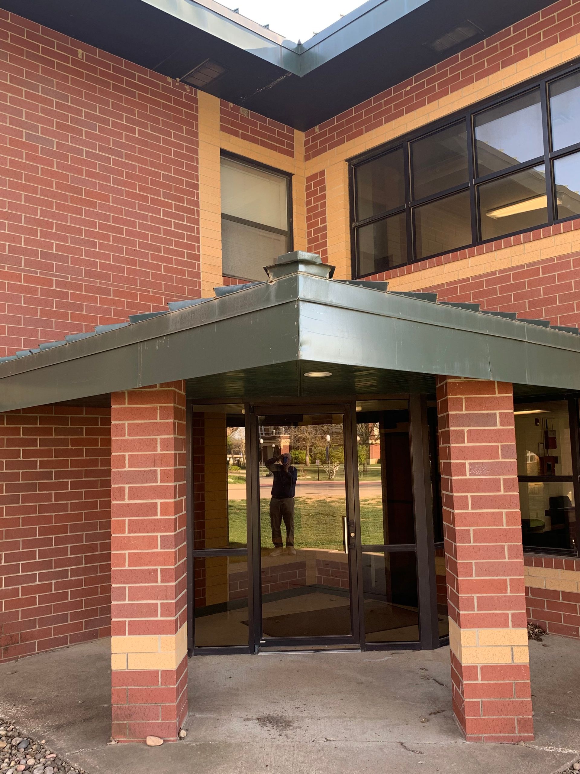 Exterior of a brick building with a covered entrance. A person is reflected in the glass doors.