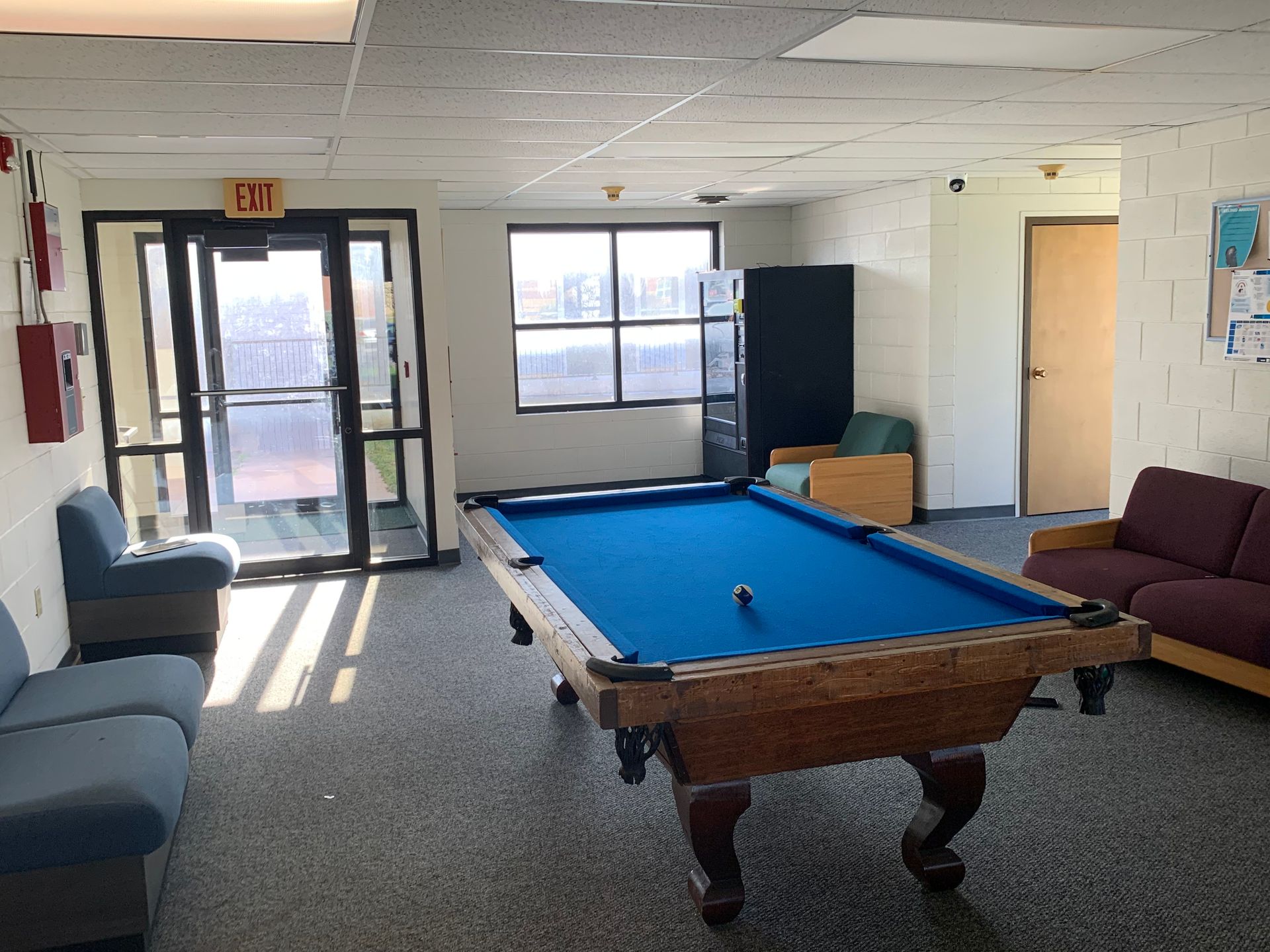 A pool table in a rec room with two blue couches, a red fire alarm, and an exit door.