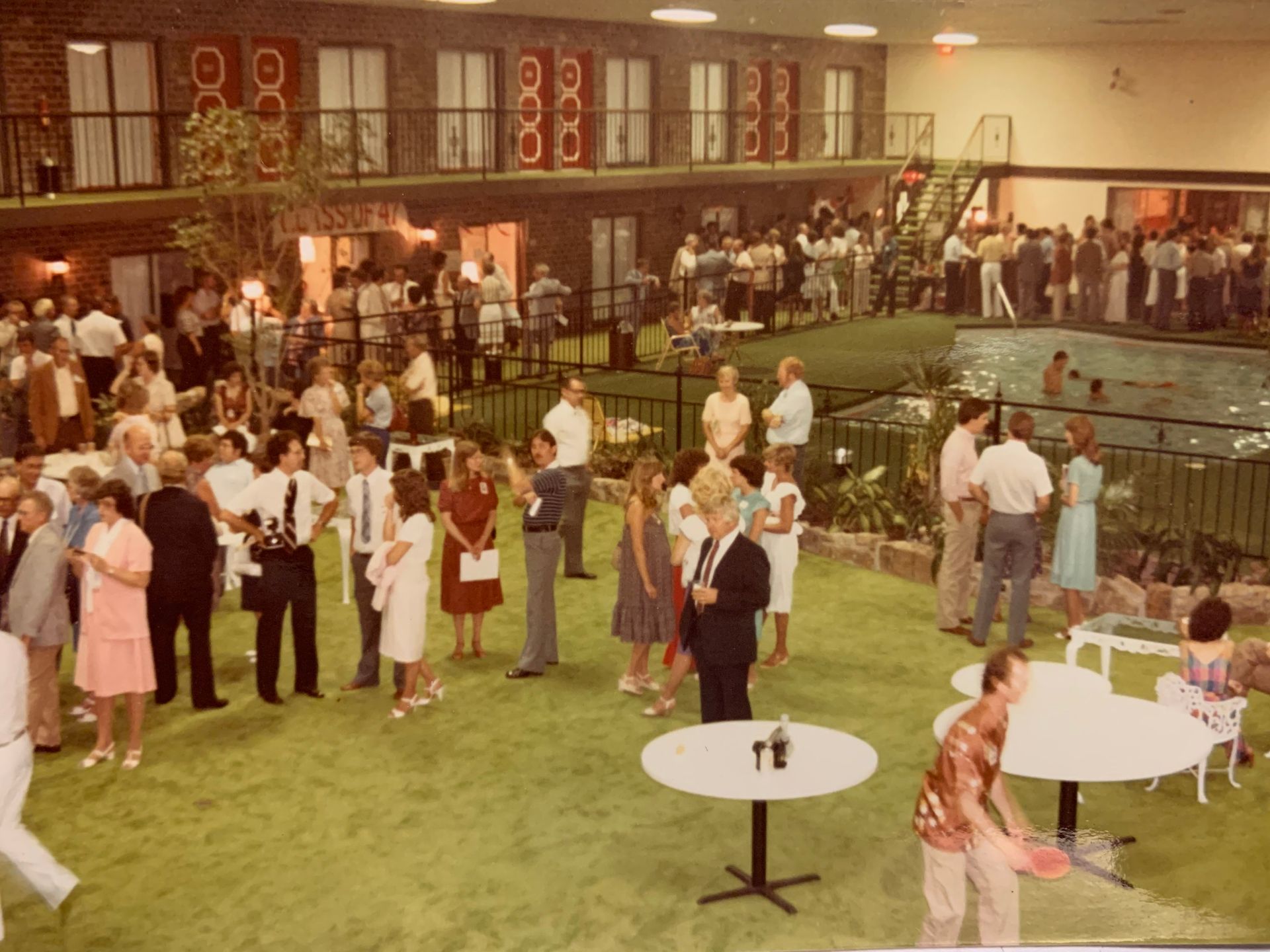 Party at indoor hotel pool, many people socializing. Green carpet, two-story building with balconies, pool in background.