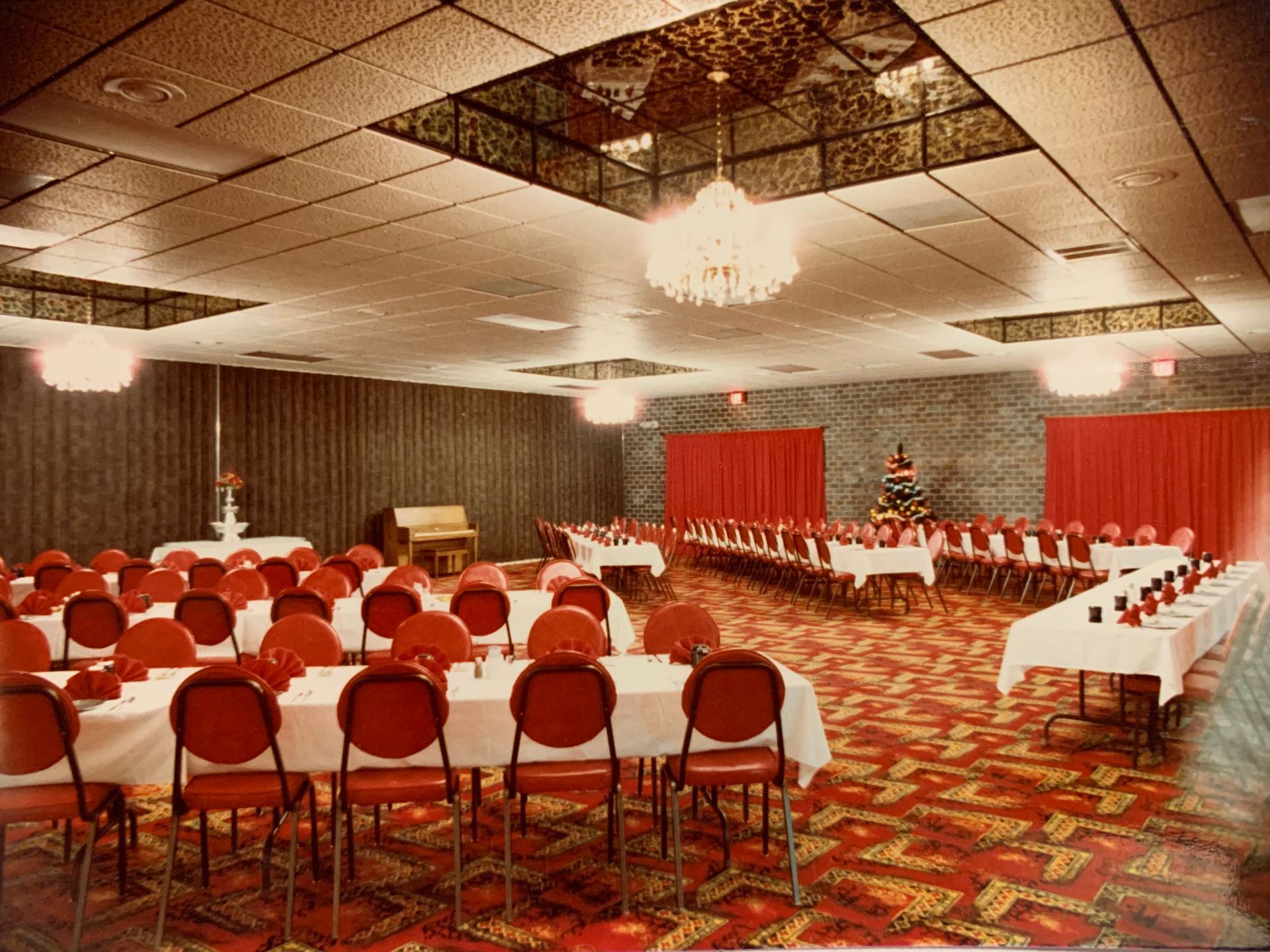 Banquet hall set for event, red chairs and tables, patterned carpet, chandelier, red curtains.