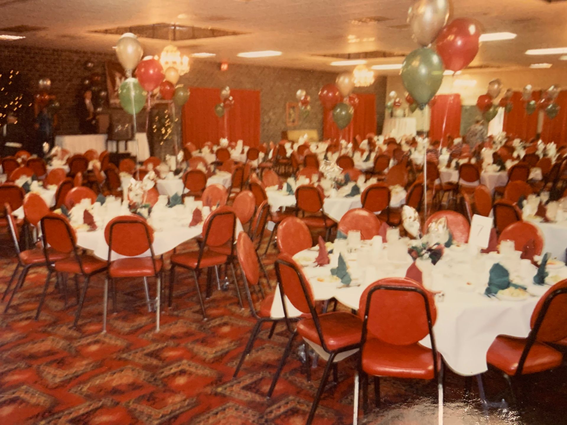 A large banquet hall with red chairs, round tables with white cloths, and balloons, ready for a Christmas celebration.