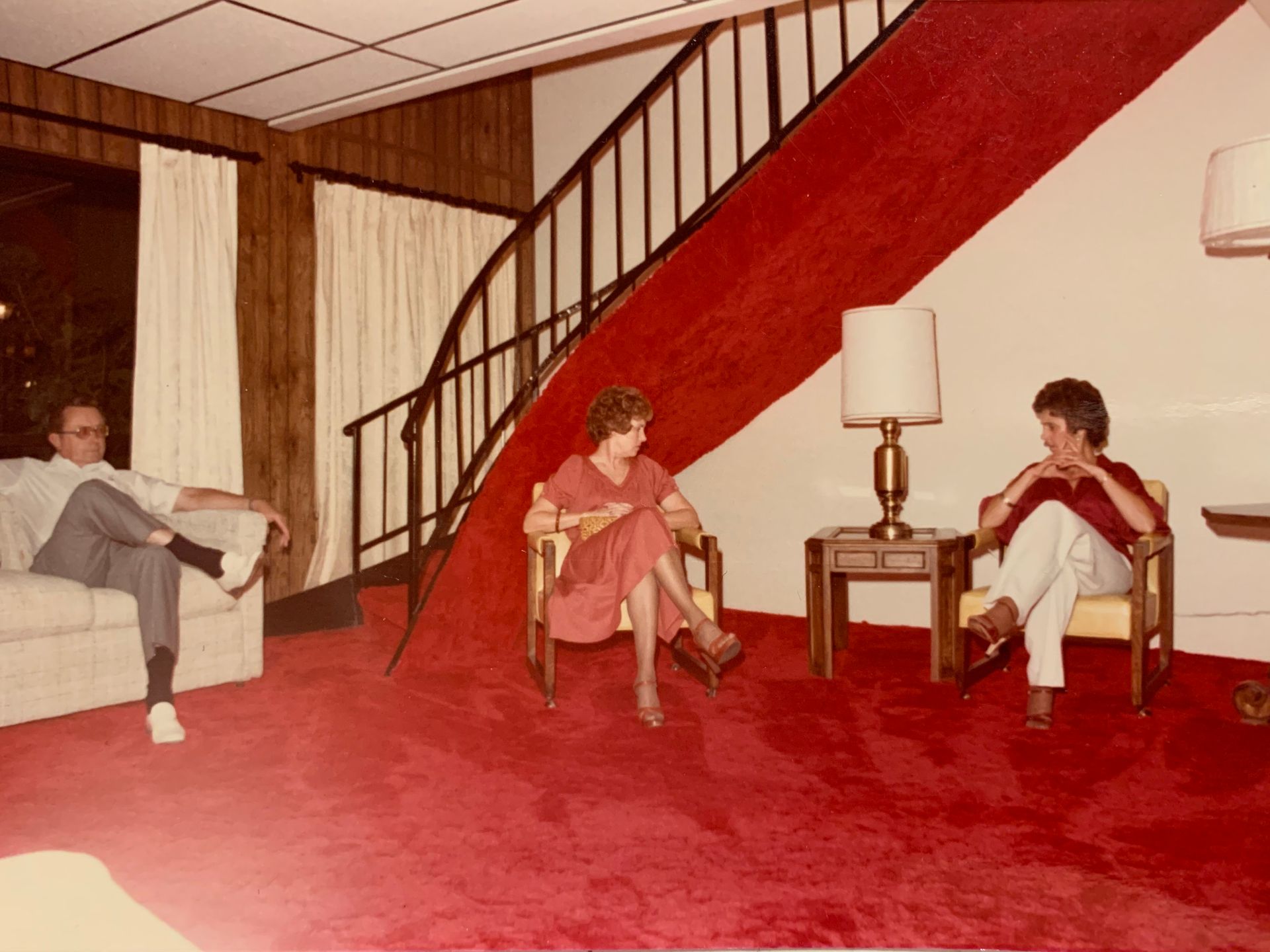 Three people seated in a room with red carpet and a staircase. One man sits on a sofa, two women in chairs.