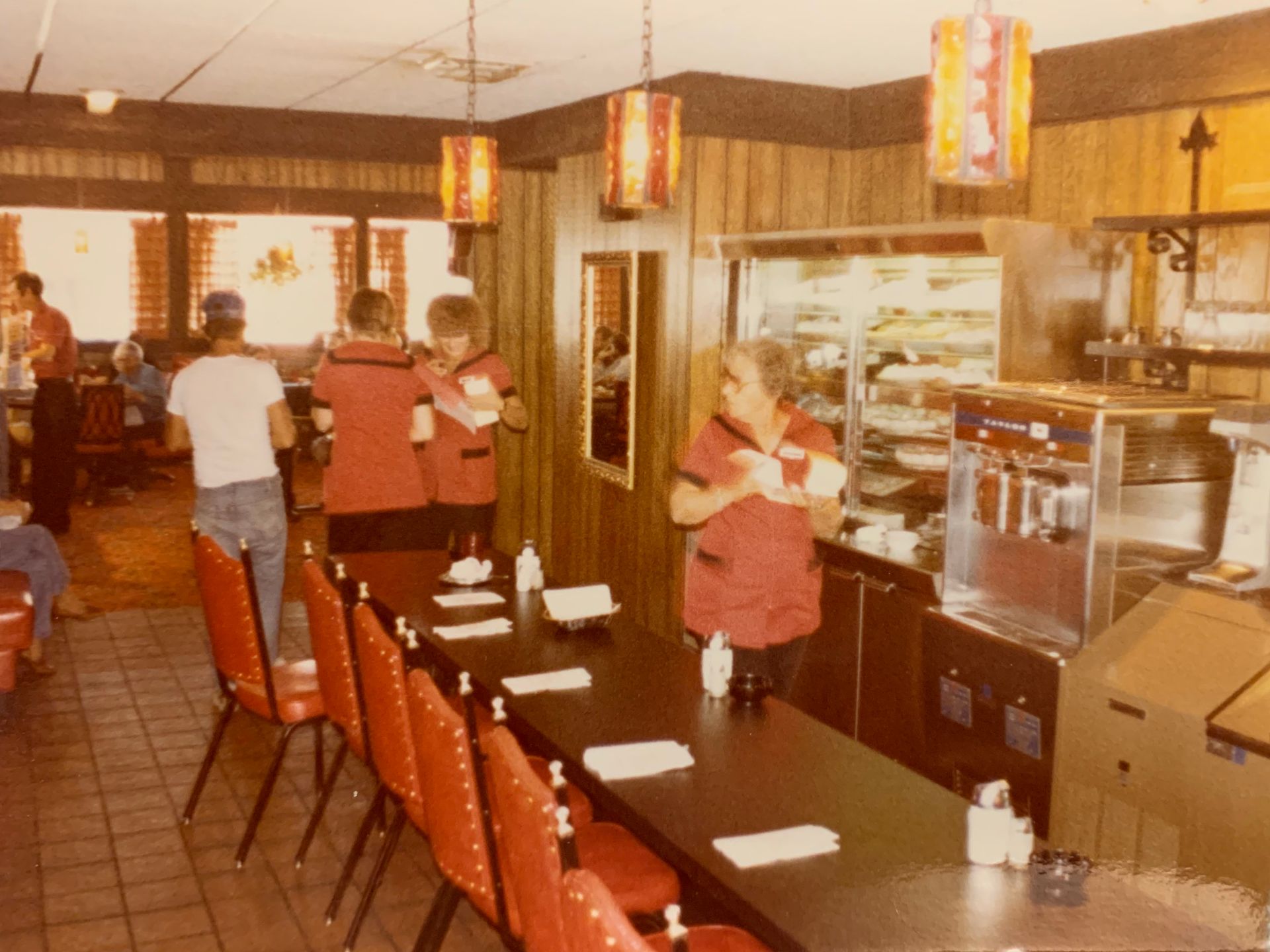 Vintage diner interior with servers in red uniforms, counter seating, and retro decor.