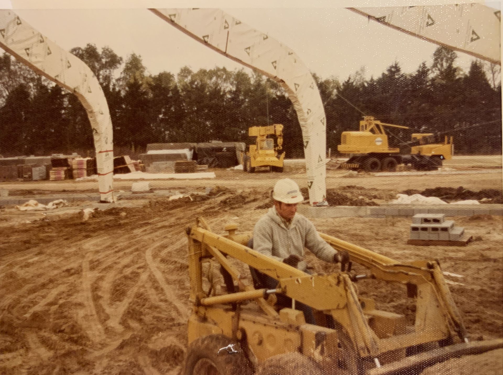 Construction worker operating a yellow skid steer loader on a dirt construction site with concrete arch supports.