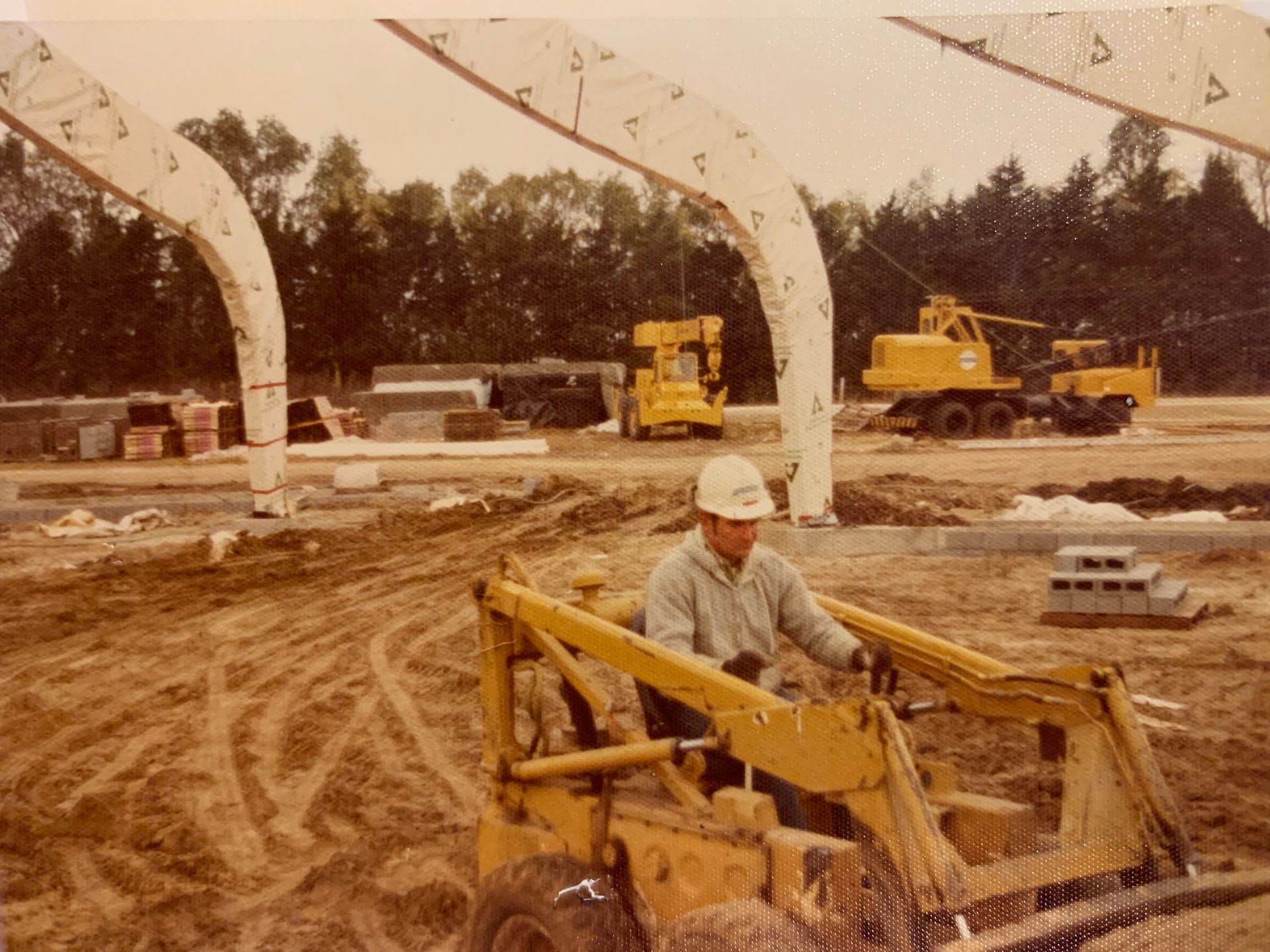 Construction site with a worker in a yellow skid steer, arched beams, and yellow construction vehicles.