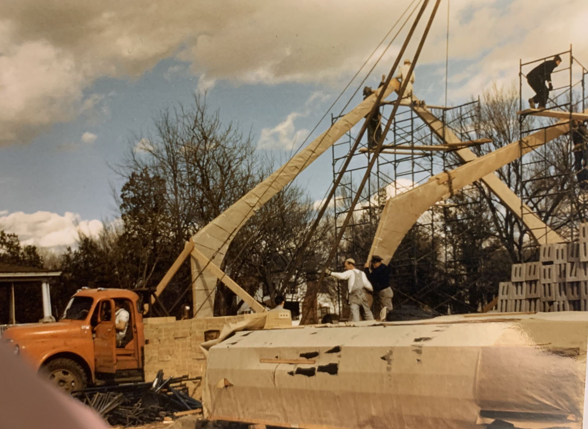 Construction site: workers erect wooden structure with a crane, orange truck, stacked stone blocks, and scaffolding.