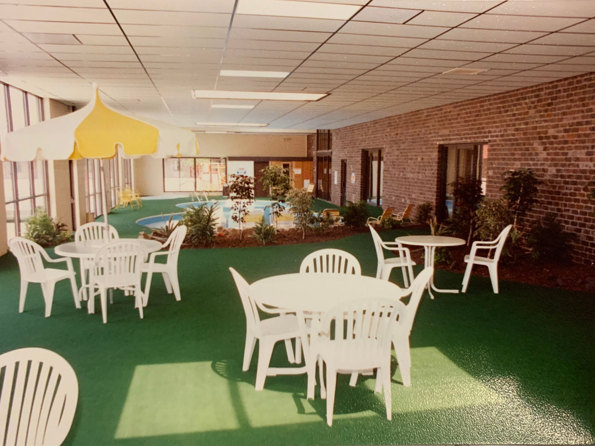 Indoor area with plastic tables and chairs, a yellow umbrella, and a small pool surrounded by plants and brick walls.