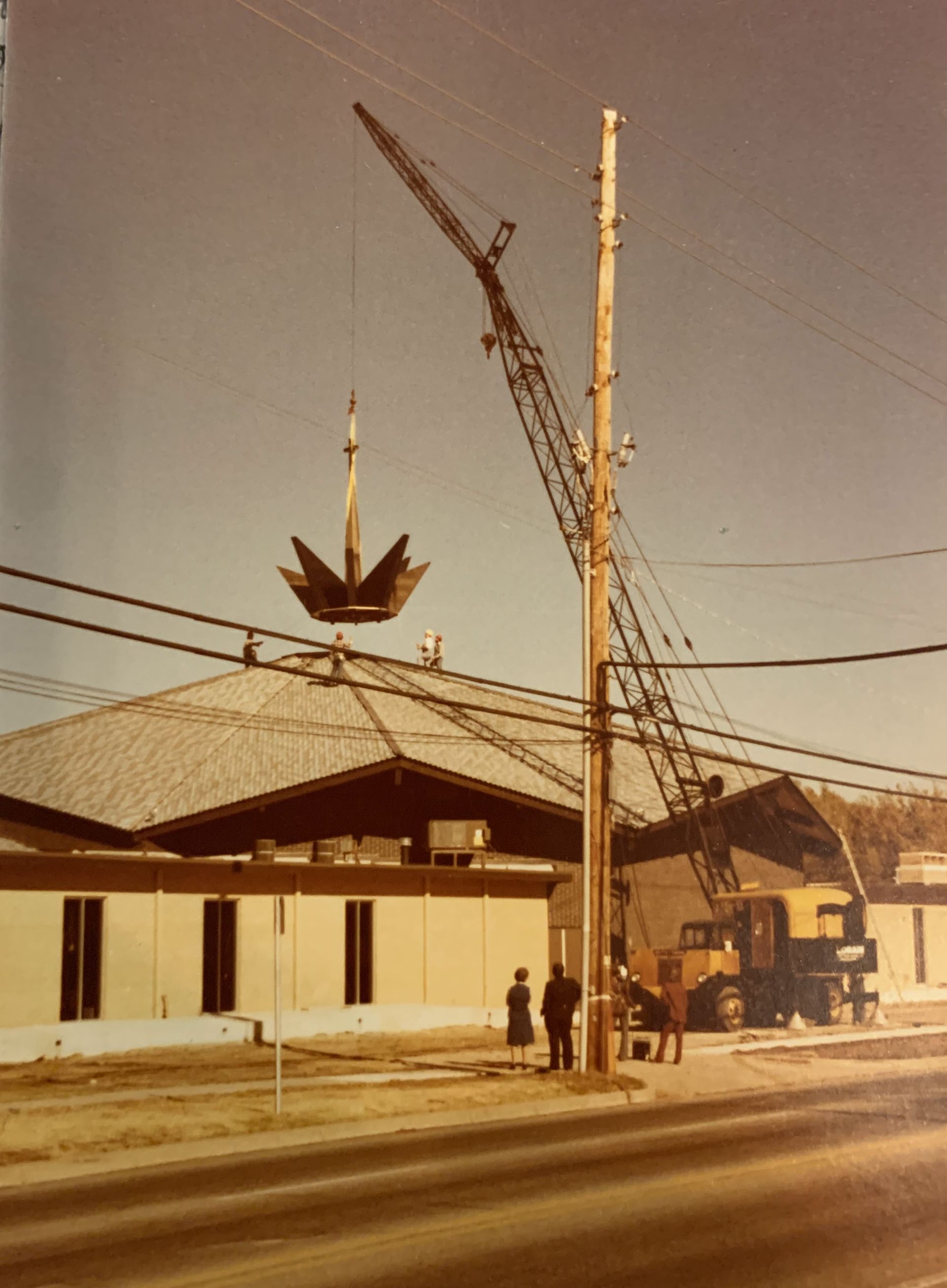 Crane lifting a geometric structure over a low building. People watch on a street with power lines.